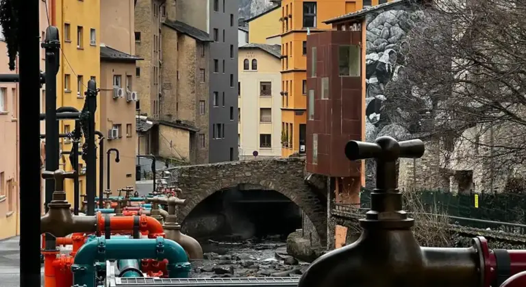 Street-level view of large red and teal water pipes with a valve wheel, set against a canal and colorful buildings in the background.