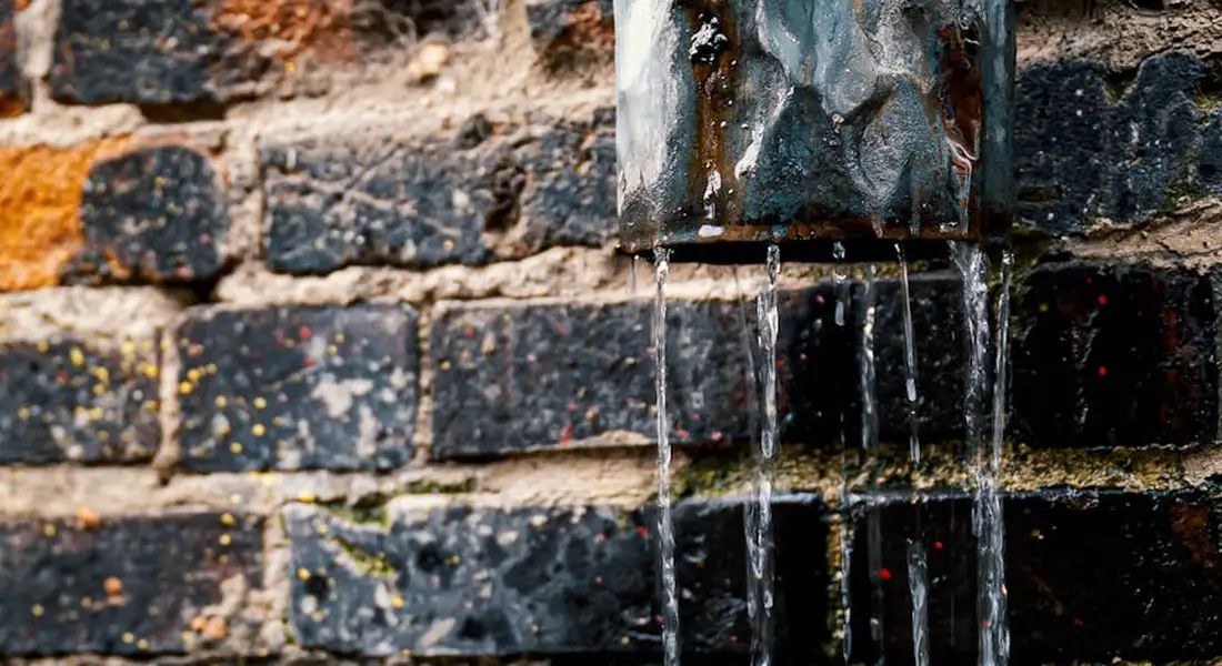 Close-up of water dripping from a metal vent pipe onto a dark brick wall, illustrating venting problems