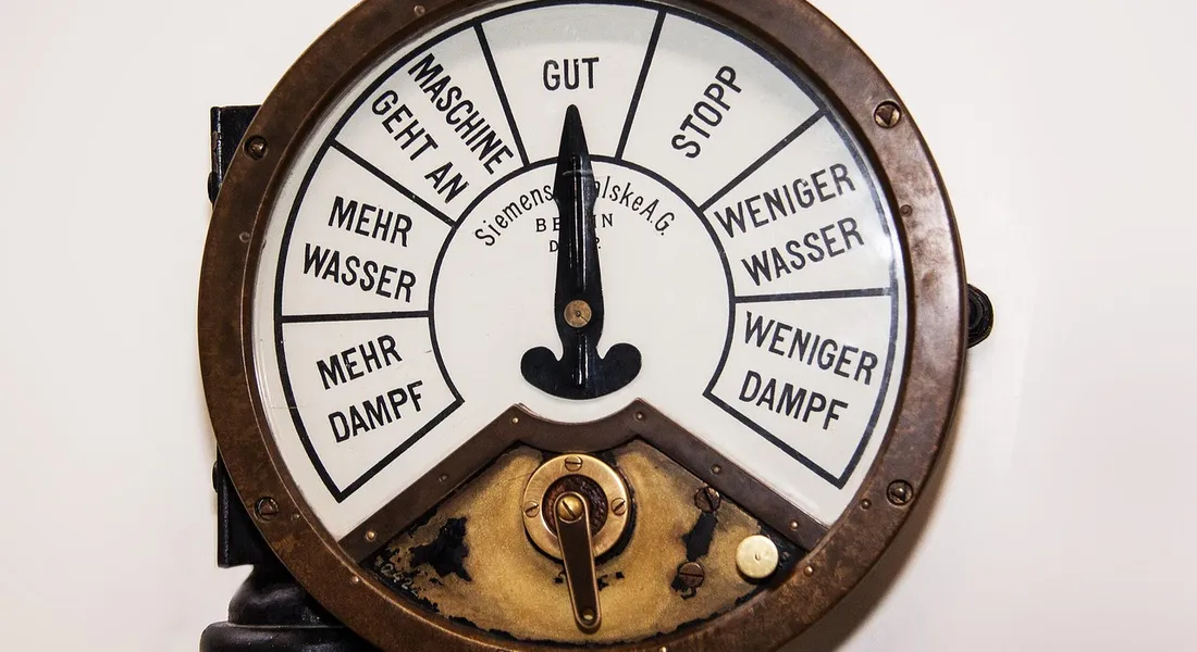 A vintage circular gauge with white segments labeled in German (Mehr Wasser, Mehr Dampf, Weniger Wasser, Weniger Dampf) indicating water and steam levels.