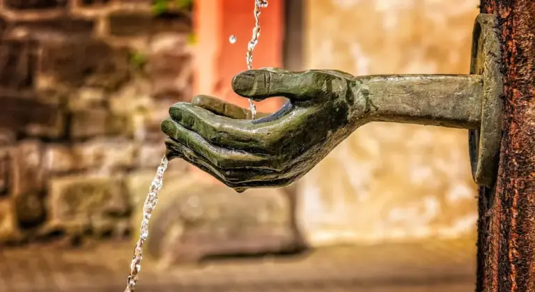 Close-up of a weathered metal hand sculpture catching a stream of water from a spout.