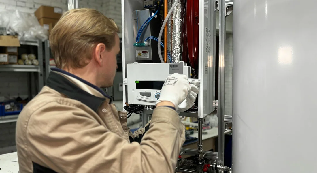 Technician wearing a tan jacket and white gloves inspects a water heater control panel in a workshop.