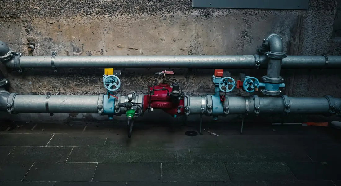Metal water pipes with blue-handled valves and a red regulator mounted along a concrete wall.