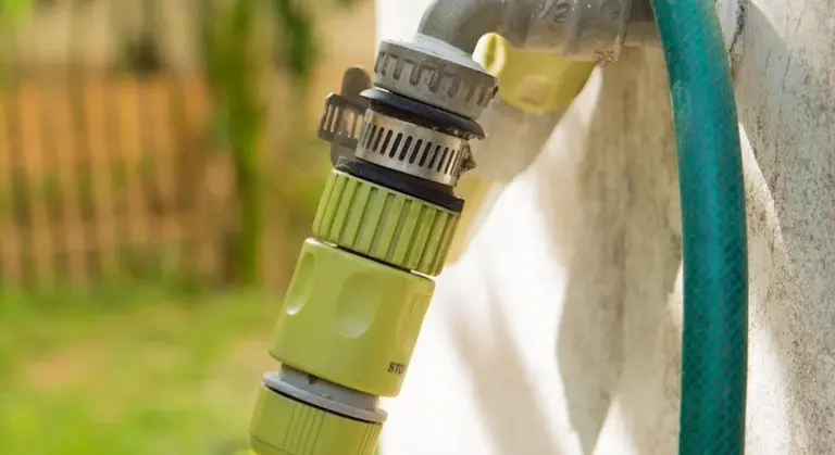 Close-up of a garden hose connector attached to an outdoor water pipe, with a green hose running beside it.