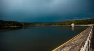 A large reservoir with a wooden railing along a dock extending toward a small white building on the far shore under dark, stormy clouds.