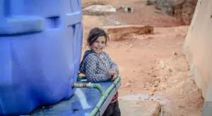 A child stands beside a large blue water storage tank with green plumbing fittings outdoors.