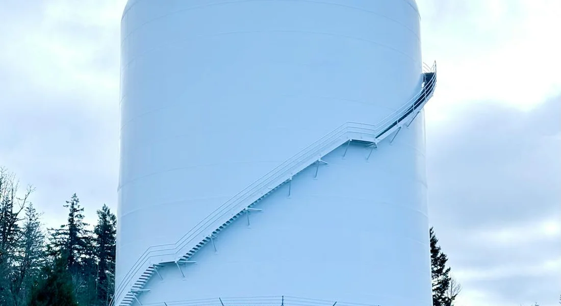 Exterior view of a tall white vertical water storage tank with an external metal staircase winding up its side, surrounded by trees under a cloudy sky.