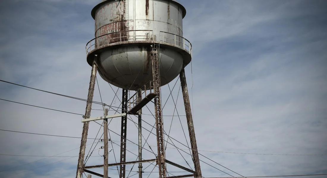 Rusty elevated metal water storage tank on a steel framework against a cloudy sky.