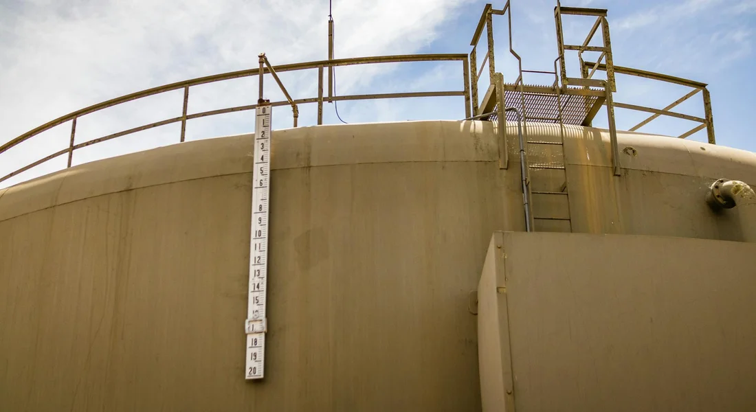 Exterior of a large beige water storage tank with a vertical measurement scale and a rooftop access ladder