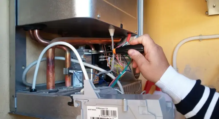 Close-up of a technician's hand examining the control panel of a water heater with exposed wires and copper pipes.