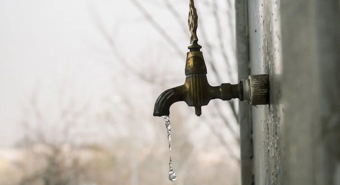 Close-up of a weathered outdoor water spigot with a small drip from the nozzle and a damp background.