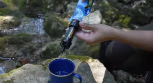 Person using a portable water purifier to fill a cup by a rocky outdoor stream