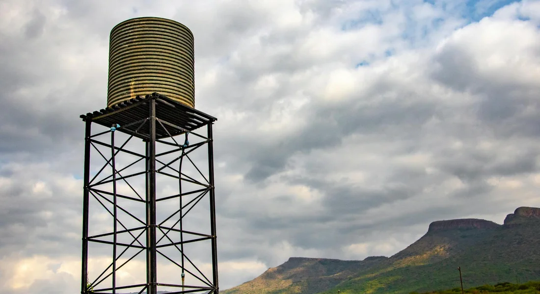 A tall cylindrical water storage tank sits atop a metal lattice tower, with a cloudy sky and distant hills in the background.