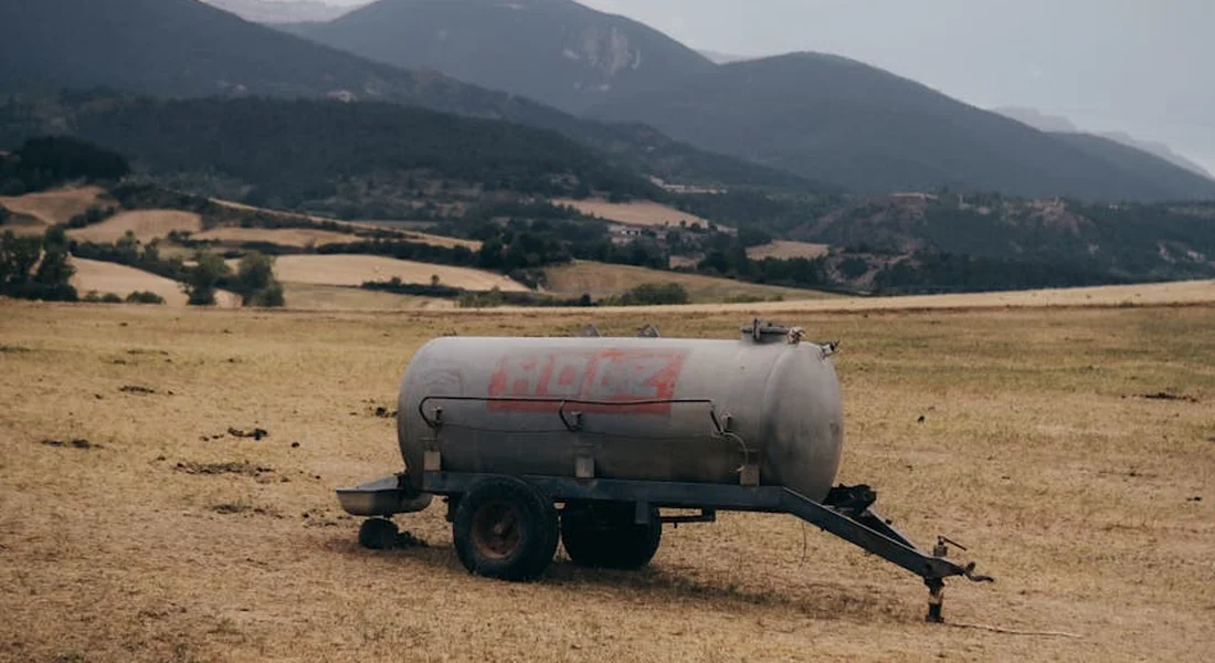 A metal water storage tank on a trailer sits in a dry field with rolling hills in the background.