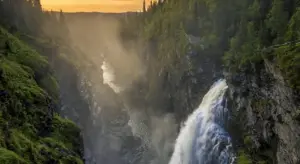 Waterfall cascading down a rocky canyon with lush green trees on both sides, at sunset.