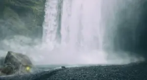 Wide scene of a powerful waterfall with mist, rocky foreground, and a dark, indistinct vehicle-like object on the left.