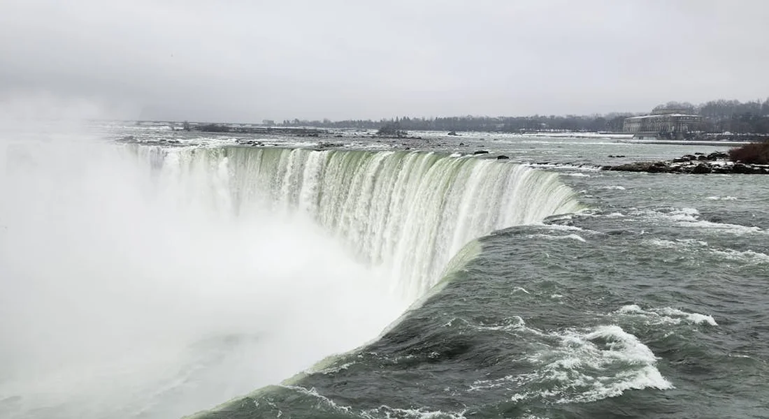Wide view of a large waterfall with mist rising into a gray overcast sky, a broad river edge visible on the far side.