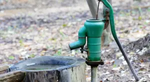 Green outdoor water pump mounted on a vertical pipe beside a tree stump in a dry, outdoor setting.