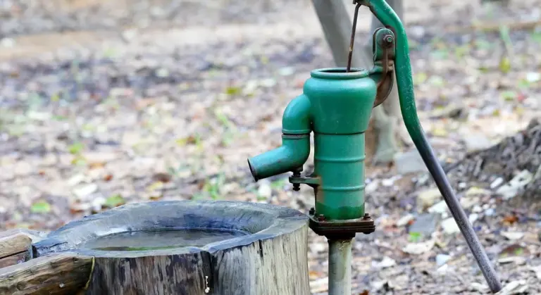 Green outdoor water pump mounted on a vertical pipe beside a tree stump in a dry, outdoor setting.
