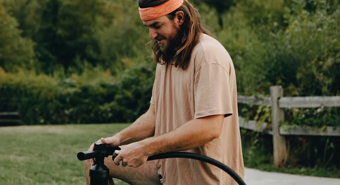 Person with long hair and an orange headband operating a hose connected to a well pump outdoors in a grassy yard.