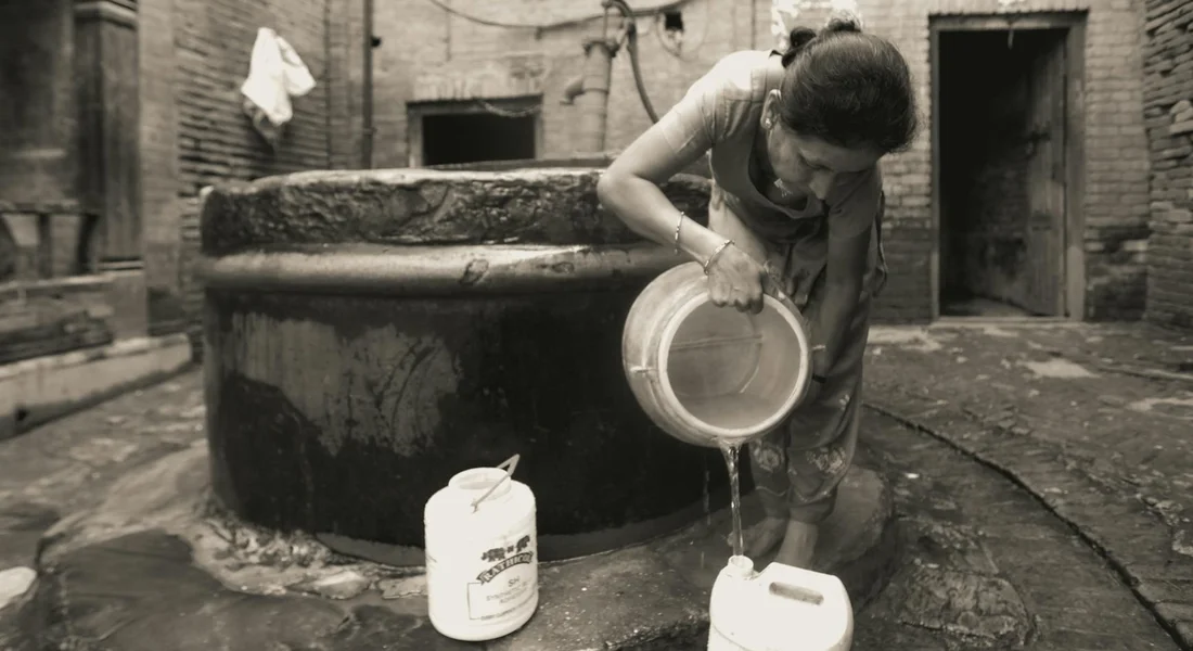 A person leans over a large circular well, pouring liquid from a jug into a smaller container, with two jugs on the damp ground nearby.