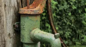 Old green metal well pump attached to a weathered wooden post with rust and greenery in the background.