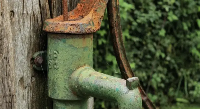 Old green metal well pump attached to a weathered wooden post with rust and greenery in the background.