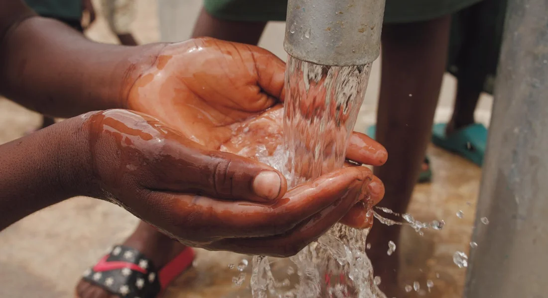 Close-up of hands cupping and rinsing under a stream of water from a well pump spigot outdoors.