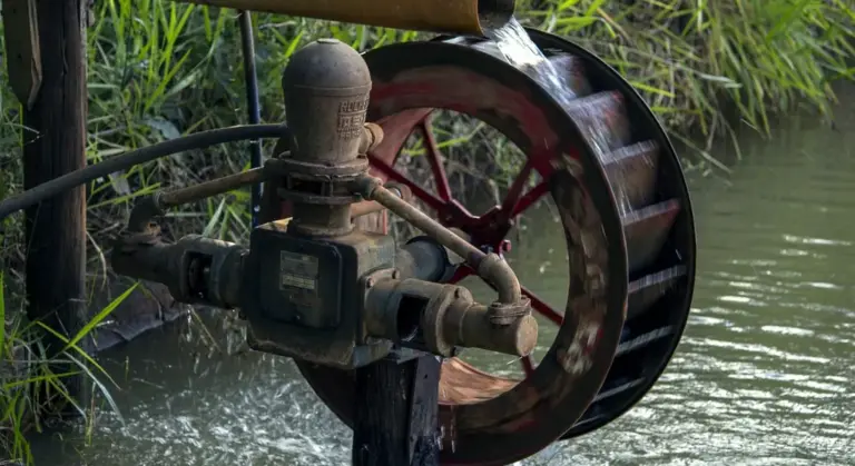 Close-up of a well pump with a red flywheel and exposed piping next to a water source.