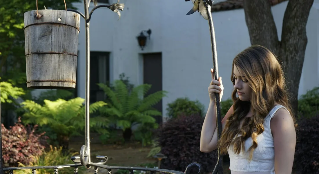 A young woman stands near an outdoor hand-powered water well pump with a wooden bucket hanging from a rope, in a garden setting.