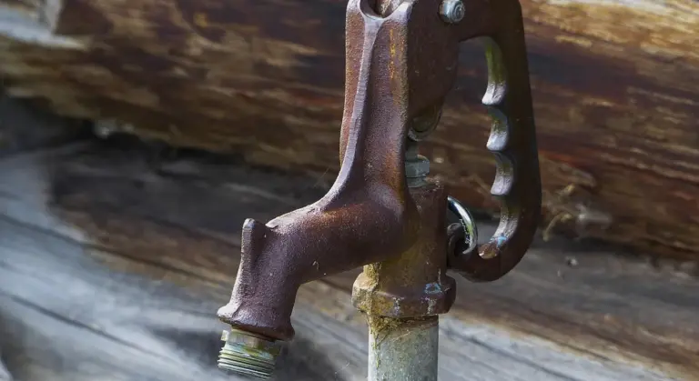 Close-up of a rusty hand-pump valve mounted on wood, representing well pump maintenance.