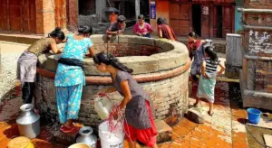 Group of people gathered around a brick well, drawing water from a communal pump in a rural setting.