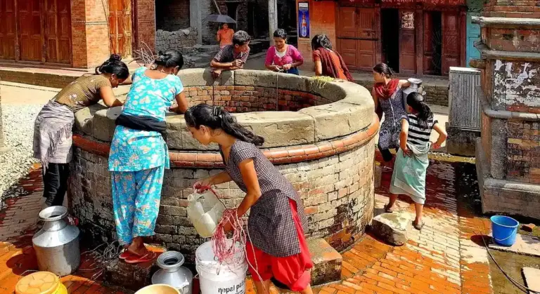 Group of people gathered around a brick well, drawing water from a communal pump in a rural setting.