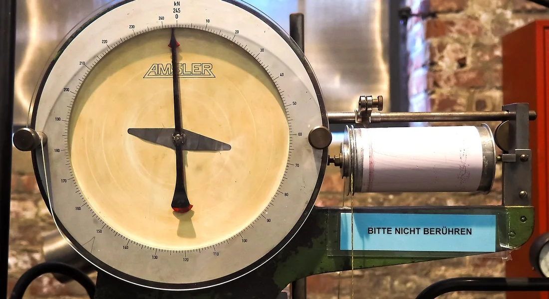 Close-up of an analog pressure gauge on a well pump with a blue warning sign in the background