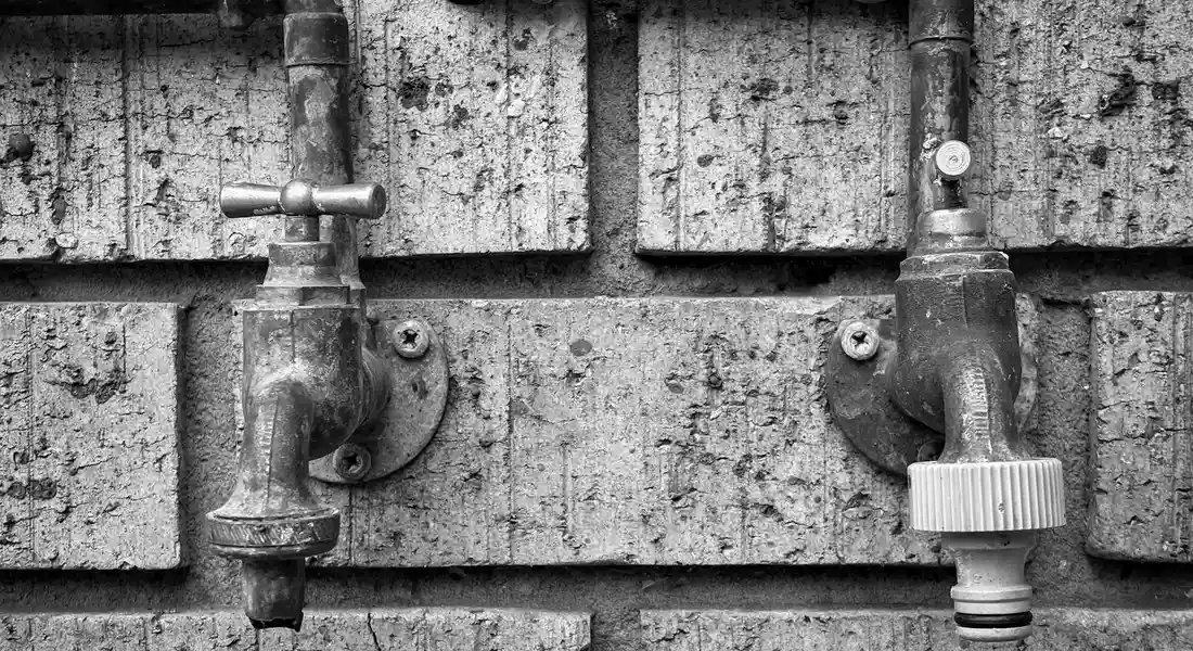 Black-and-white photo of outdoor pool plumbing on a brick wall, featuring valve handles and piping.