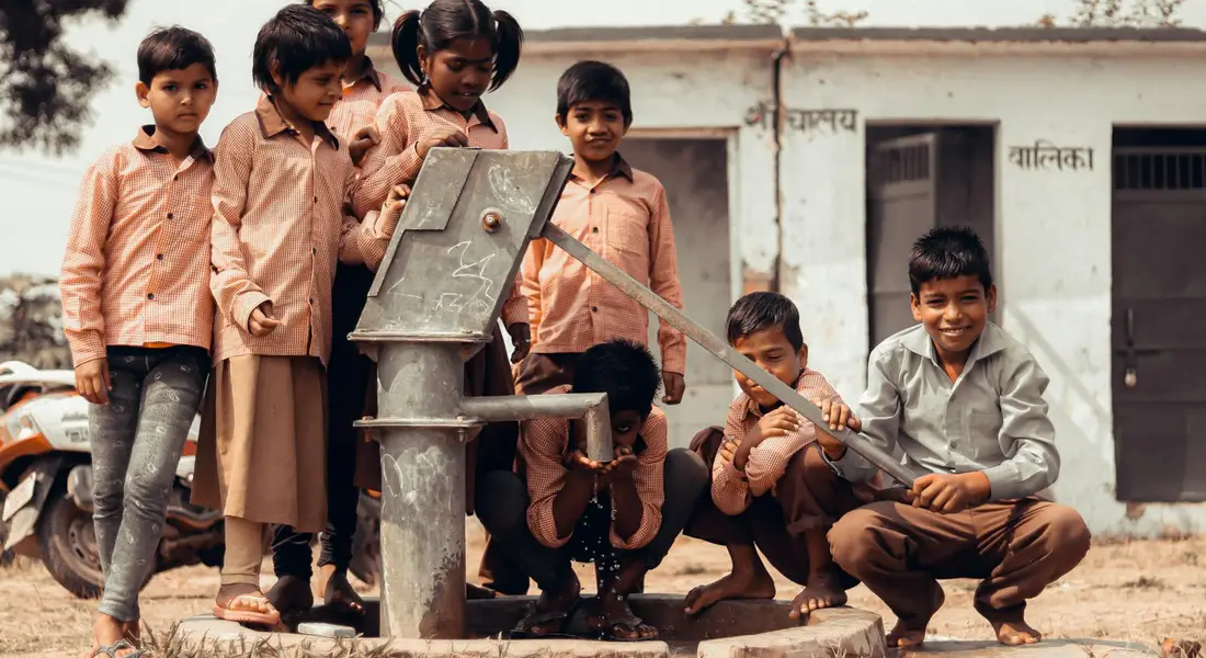 Group of children gathered around a manual hand water pump outdoors.