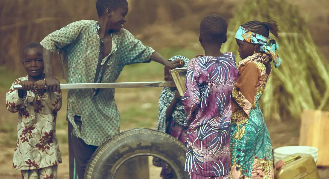A family stands around a hand-operated water pump in a rural setting, with a man operating the pump and children nearby in colorful clothing.